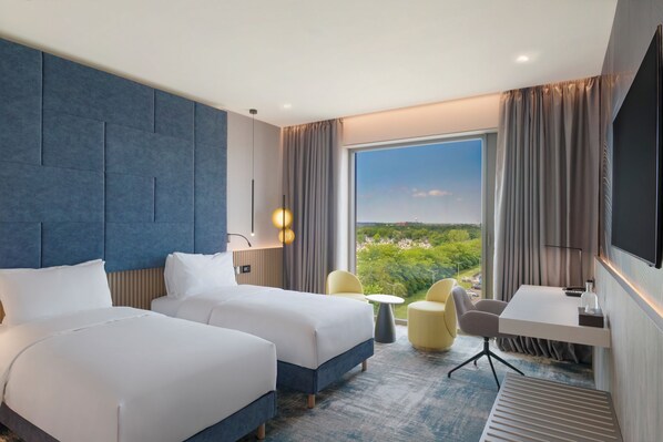Habitación de hotel con dos camas, un escritorio, una silla, un televisor y un ventanal con vistas.