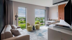 Ein modernes Hotelzimmer mit großem Fenster, einer Couch, zwei Sesseln, einem kleinen Tisch und einem Schreibtisch mit Stuhl.