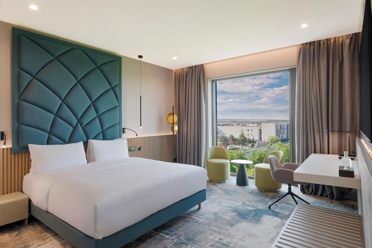 Ein modernes Hotelzimmer mit einem großen Bett, einem Schreibtisch, einem Sessel und Blick auf die Stadt.