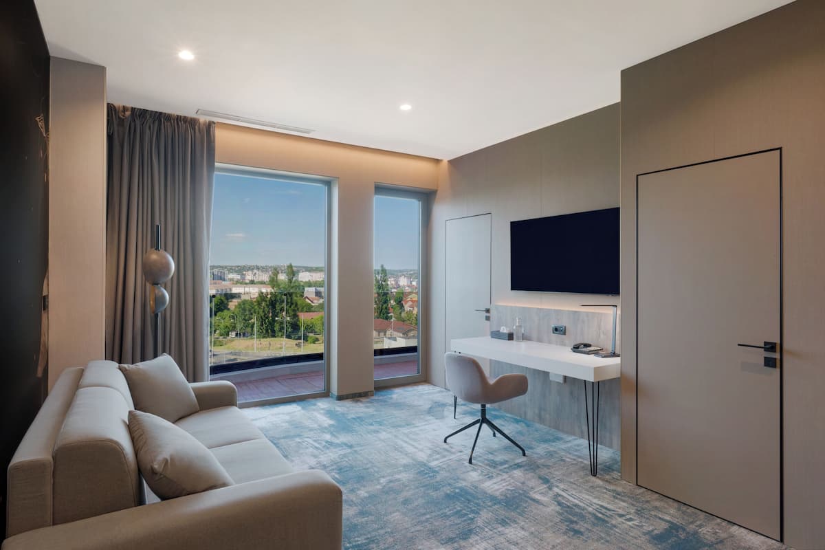 Ein modernes Hotelzimmer mit großem Fenster, einer Couch, einem Schreibtisch und einem Flachbildfernseher.