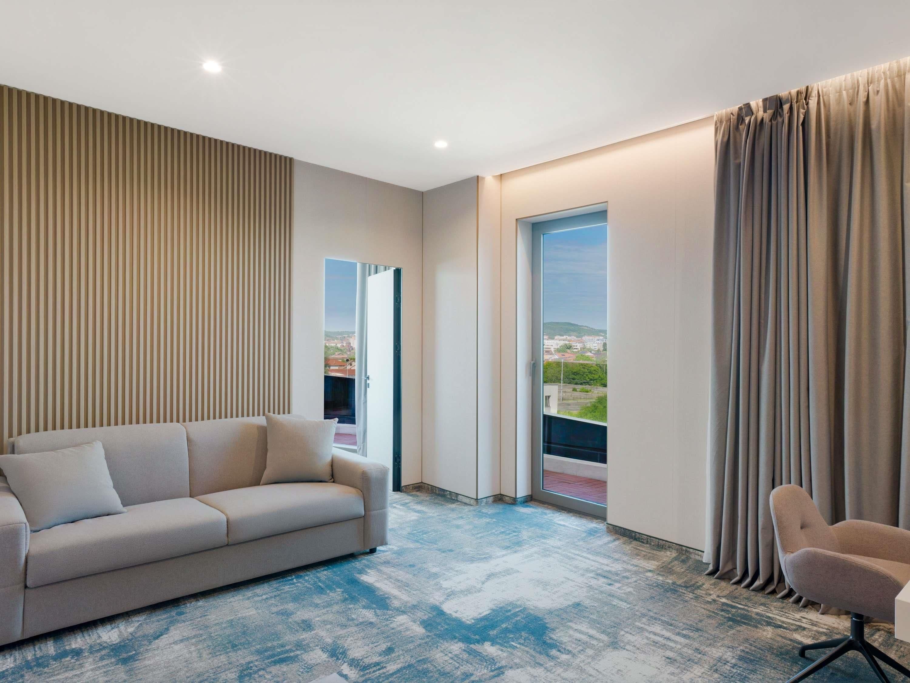 A modern hotel room with a sofa, a chair, a large window with curtains, and a view of the cityscape.