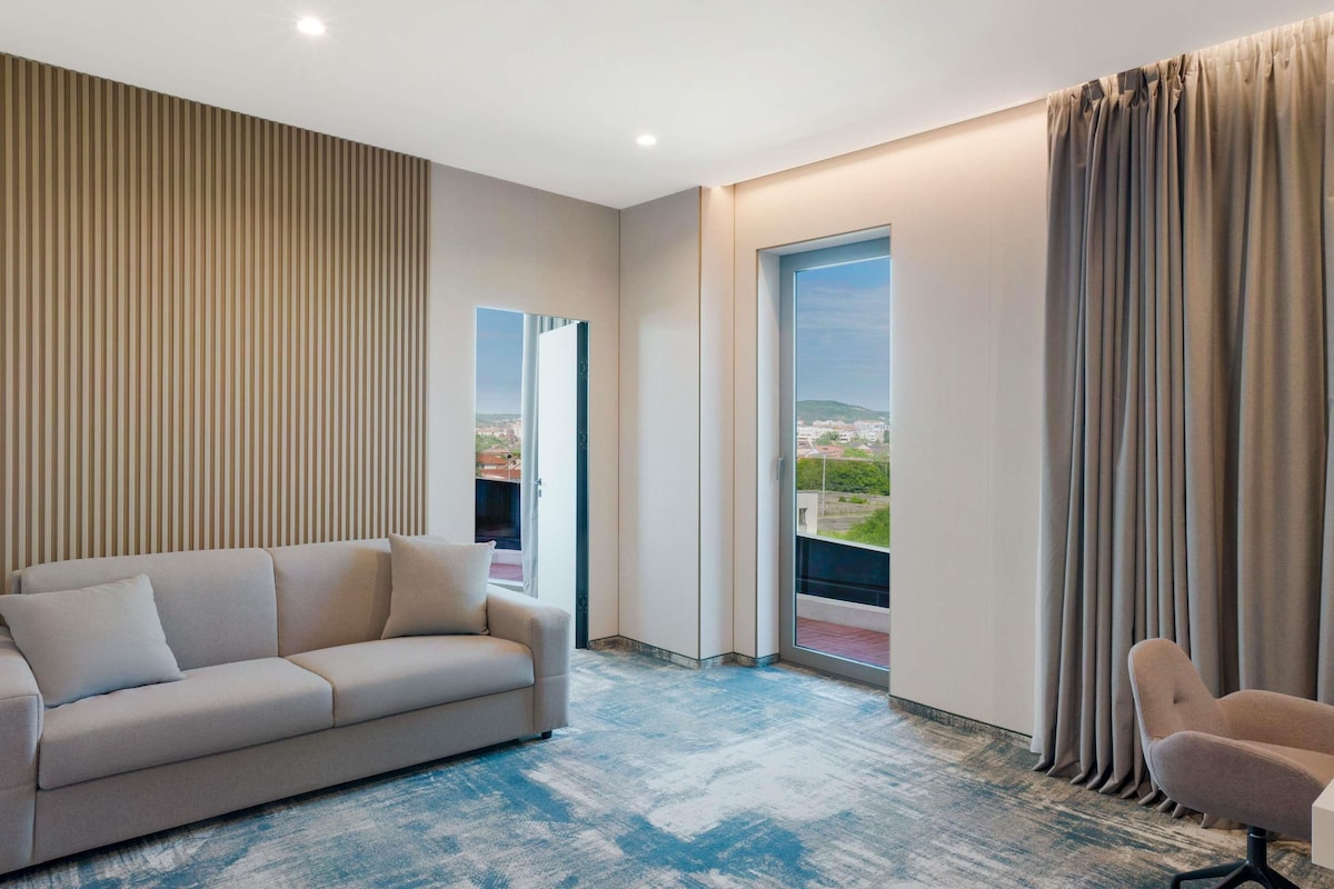 Una habitación de hotel moderna con un sofá, una silla, un ventanal con cortinas y vistas a la ciudad.