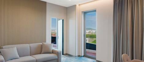 Una habitación de hotel moderna con un sofá, una silla, un ventanal con cortinas y vistas a la ciudad.