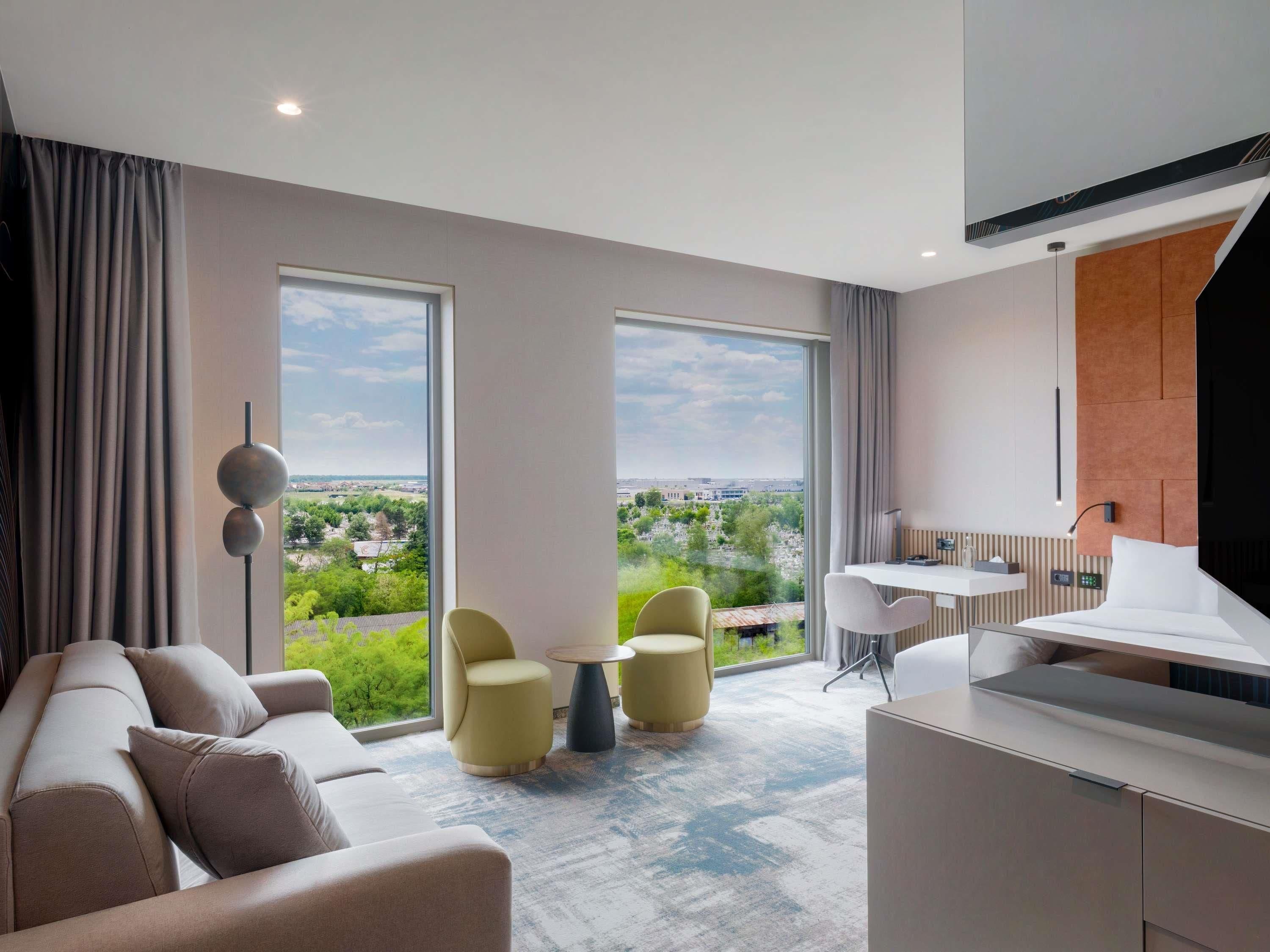 Una habitación de hotel moderna con un ventanal grande, un sofá, dos sillones, una mesita, un escritorio y una cama.