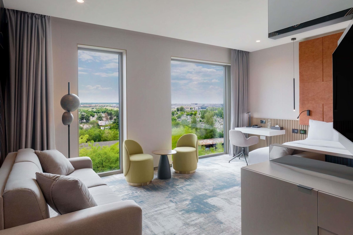 Una habitación de hotel moderna con un ventanal grande, un sofá, dos sillones, una mesita, un escritorio y una cama.