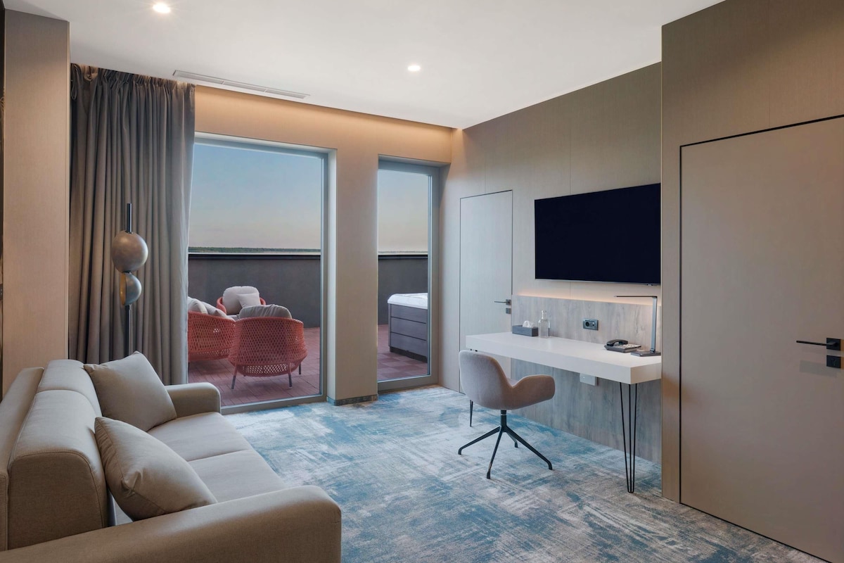 Habitación de hotel con sofá, escritorio con silla, televisor y balcón con vistas.