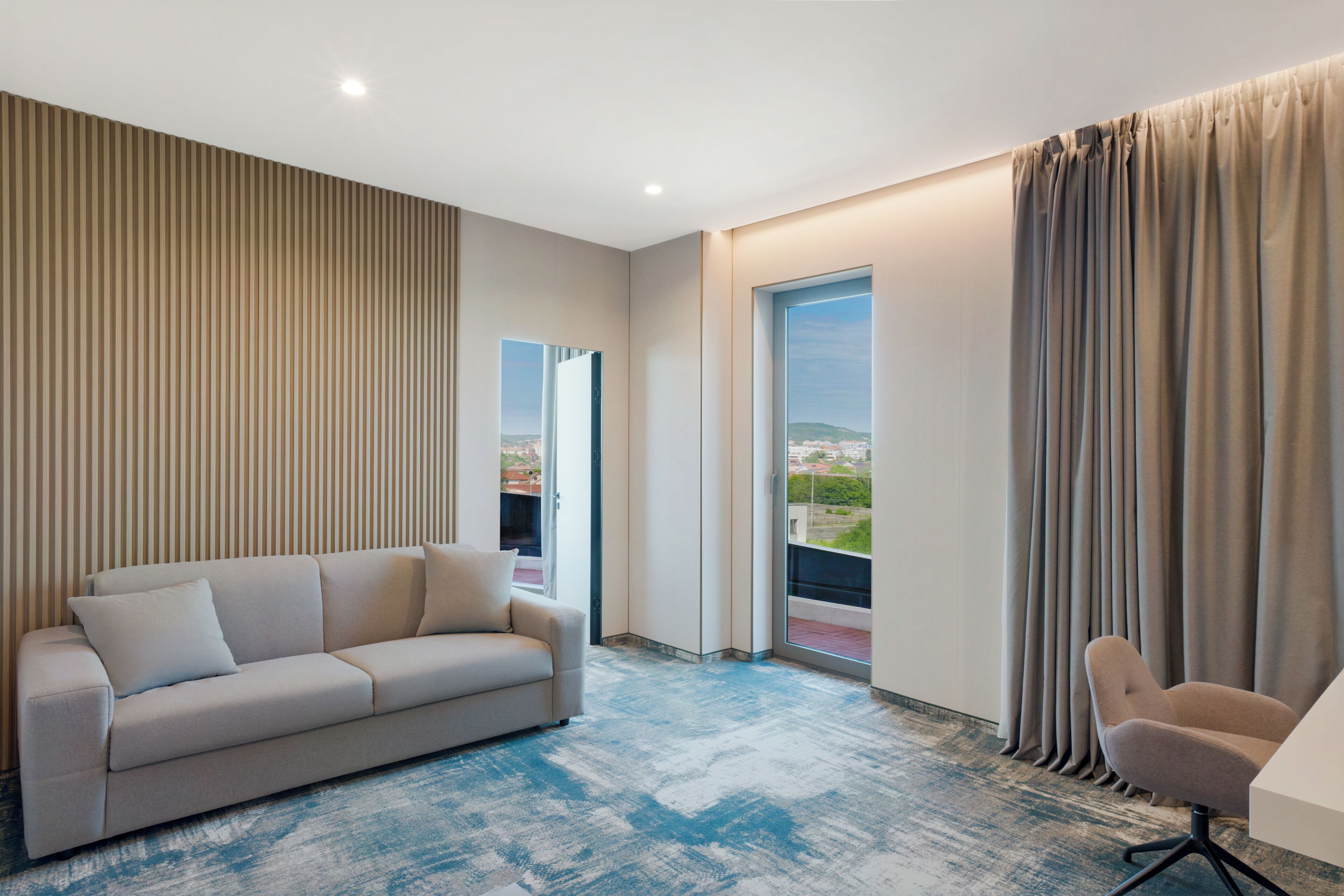 A modern hotel room with a sofa, a chair, a large window, and a view of the city.