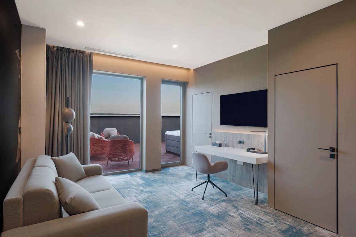 Ein modernes Hotelzimmer mit Sofa, Schreibtisch, Stuhl, Fernseher und einem Balkon mit Blick.