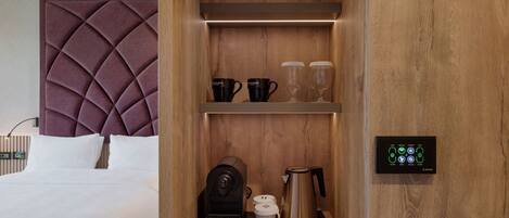 A hotel room with a tufted headboard, a coffee maker, mugs, a glass, and a control panel.