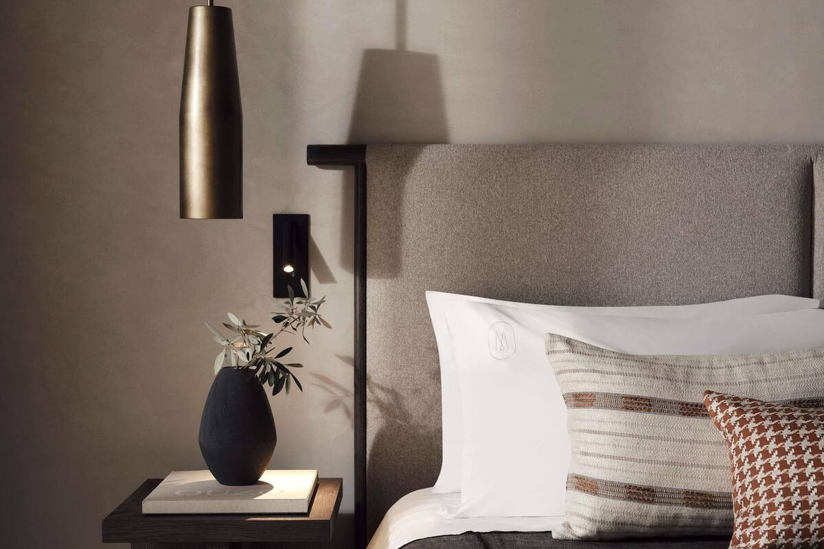 En pænt redt seng med puder, et natbord med en vase og en bog, samt en lampe ophængt på væggen.