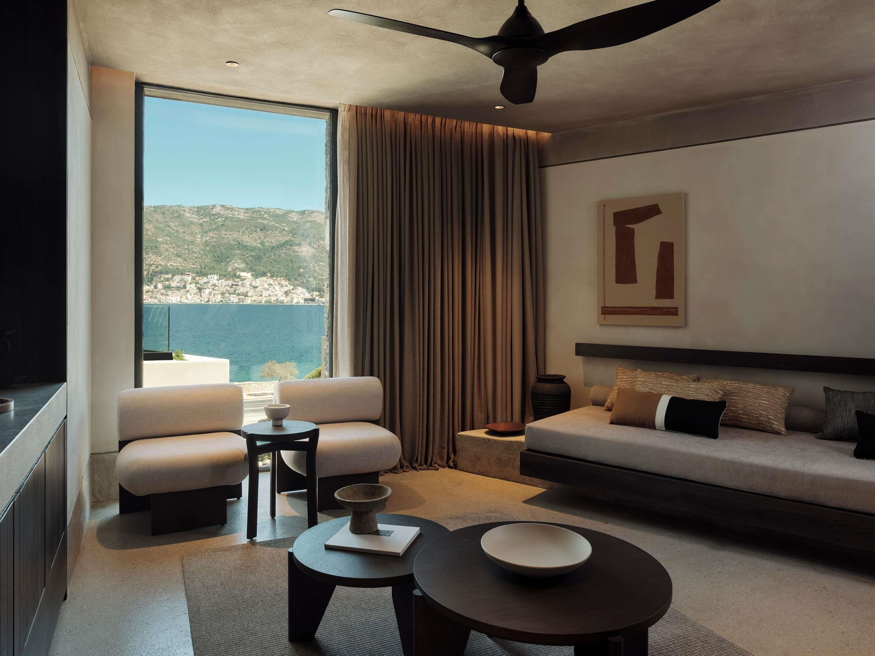 Une chambre d’hôtel moderne dotée d’une grande fenêtre offrant une vue sur la mer et les collines, d’un canapé, d’une table à manger et d’un ventilateur de plafond.