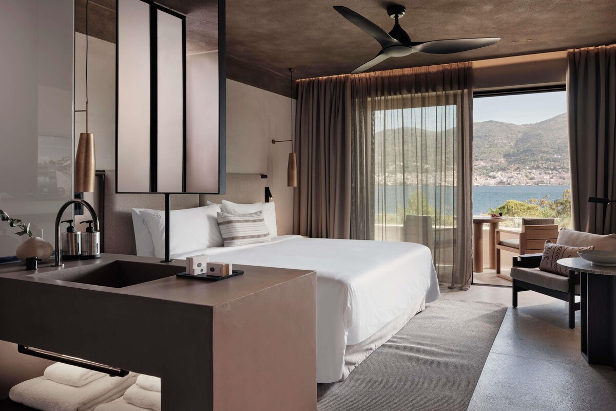 Habitación de hotel moderna con una cama grande, baño con lavamanos y un balcón con vista al mar.