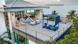 Una casa moderna con balcón que da a una masa de agua, amueblada con asientos exteriores y una silla colgante.