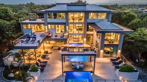 Una casa moderna de varios pisos con piscina, mobiliario exterior y un amplio frente de cristal.