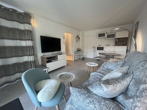 A living room with a sofa, a TV, a dining table, and a kitchen area. - Port Fréjus, Le Cesaree, Exceptional 2-bedroom apartment with garden and pool views, ideal for 4 lucky guests, air conditioning and garage (Fréjus)