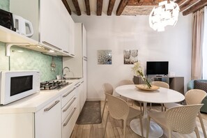 A modern kitchen with white cabinets, a microwave, and a gas stove. There is a dining area with a round table and wicker chairs. - Domus Rialto Emerald- Bright 2 Bedrooms in Central (Venice)