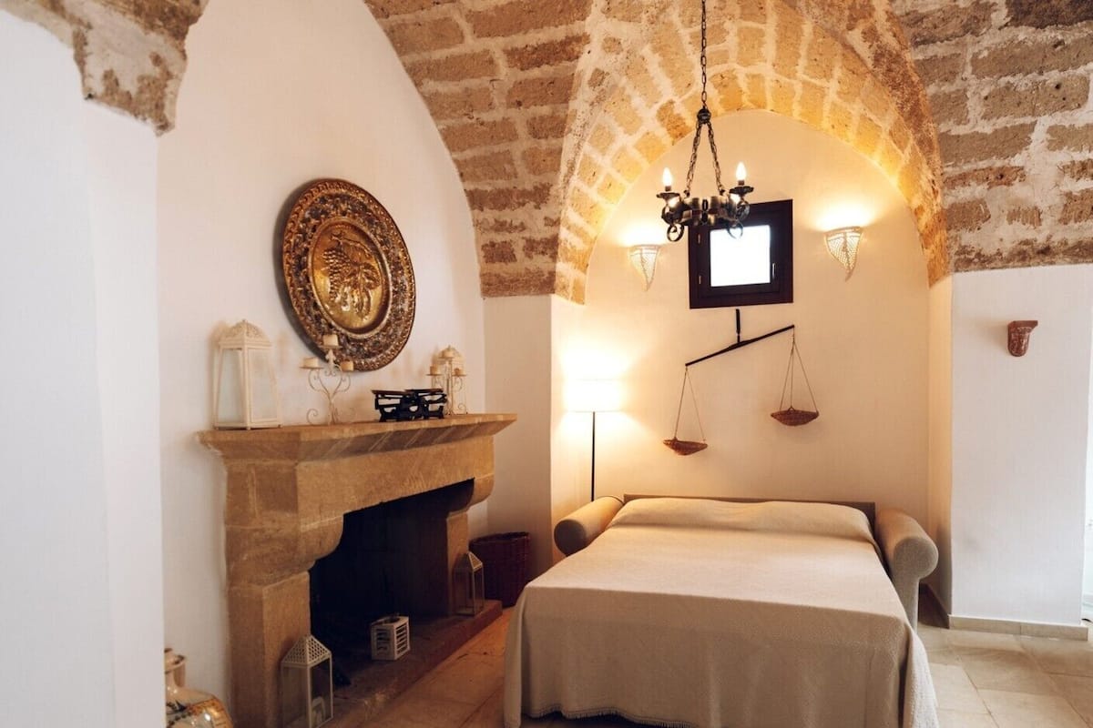 A room with a stone fireplace, a bed with a beige bedspread, a chandelier, and a wall-mounted scale.