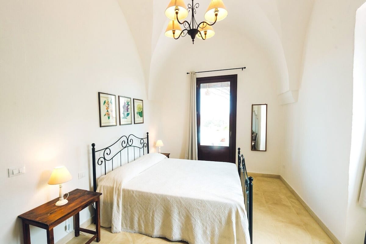 A bedroom with a bed, a nightstand, a mirror, a chandelier, and framed artwork on the wall.