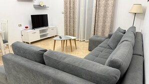 Ett modernt vardagsrum med en grå soffa, ett litet vitt bord, en tv på ett vitt skåp och en lampa.