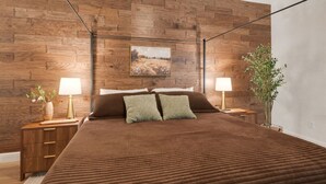 A bedroom with a wooden wall, a bed with a brown bedspread, two bedside tables with lamps, and a plant. - Emily House | 3 King Beds | Pet Friendly (Ada)