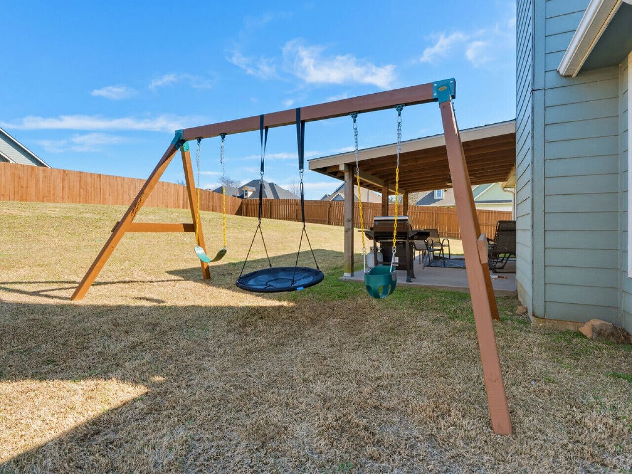 Un parque infantil de madera con columpio y trampolín, un porche cubierto y una casa al fondo.