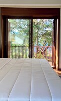 Una habitación con cama, un balcón con barandilla roja y vistas a árboles y una casa.