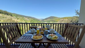 Ein Balkontisch mit Frühstücksangebot, Blick auf grüne Hügel und einen klaren Himmel.