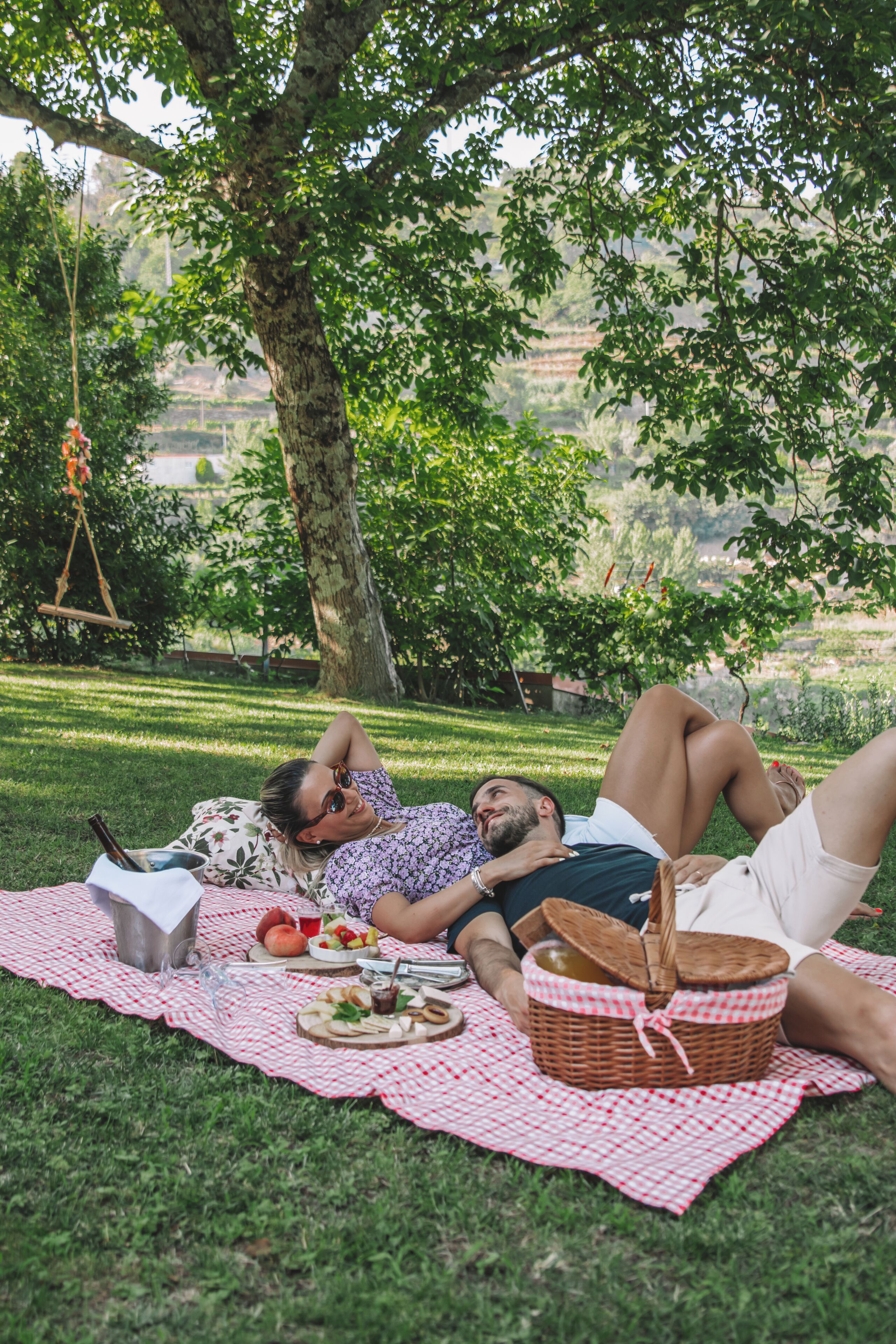 A picnic setup on a grassy area with a checkered blanket, a picnic basket, and a metal container.