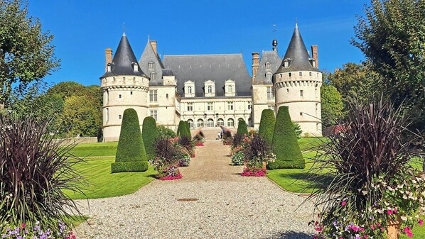 A large, historic castle with a symmetrical facade, multiple towers, and a well-maintained garden with hedges and flower beds.