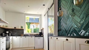 A modern kitchen with white cabinets, a dishwasher, and a window with a view of a house. - “Le Secret des Cordelières” Cottage – Charming Character Residence in Normandy (Neufchâtel-en-Bray)