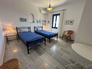 A bedroom with two beds, a chair, a table, and a window with curtains. - Villa Drionisi in Tzanes, Paros · 6 Guests (Tzanes)