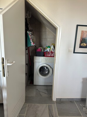 A laundry room with a washing machine, a red laundry basket, and a shelf with cleaning supplies. - Villa Drionisi in Tzanes, Paros · 6 Guests (Tzanes)
