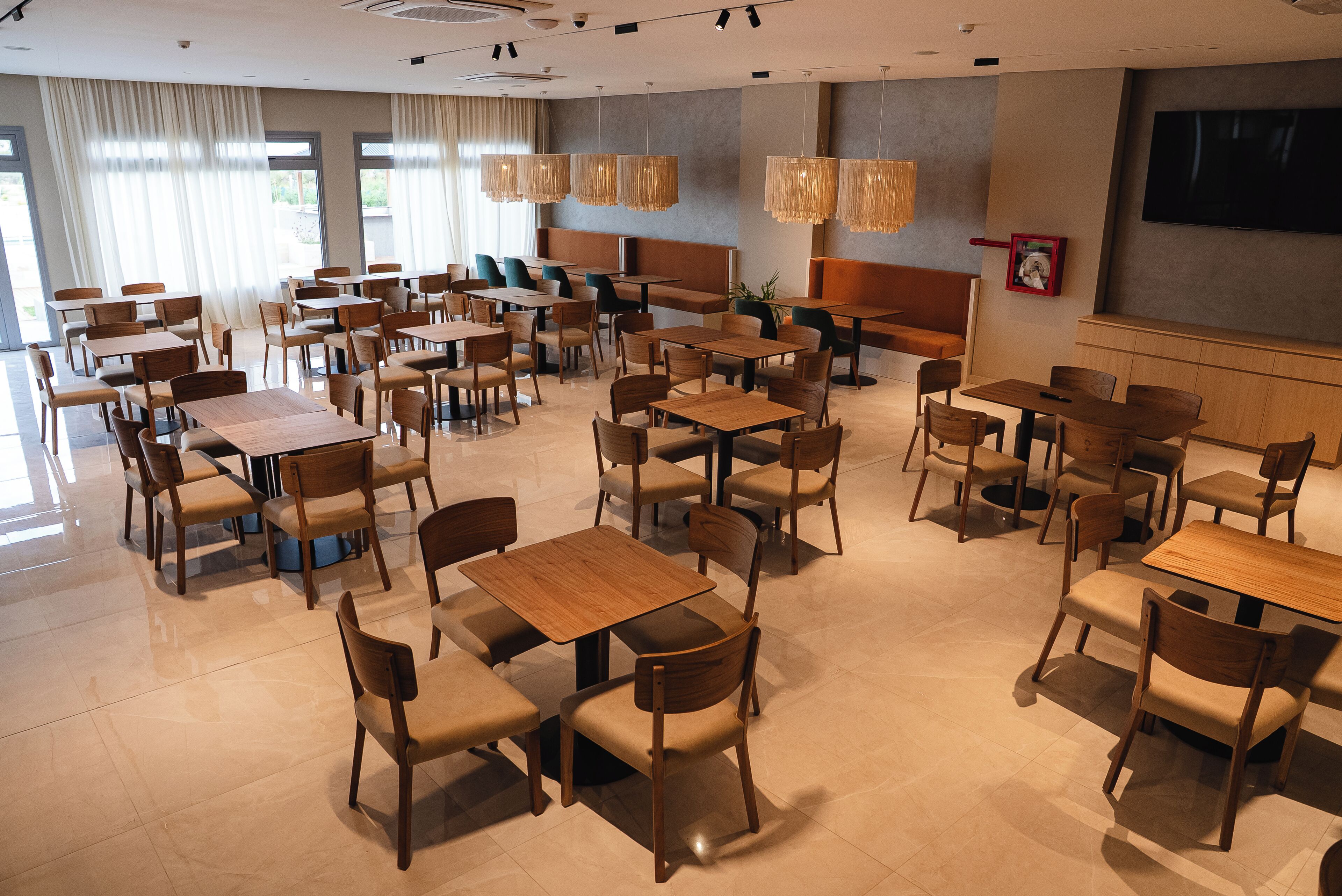 A spacious dining area with multiple tables and chairs, large windows with curtains, and a flat-screen TV mounted on the wall.