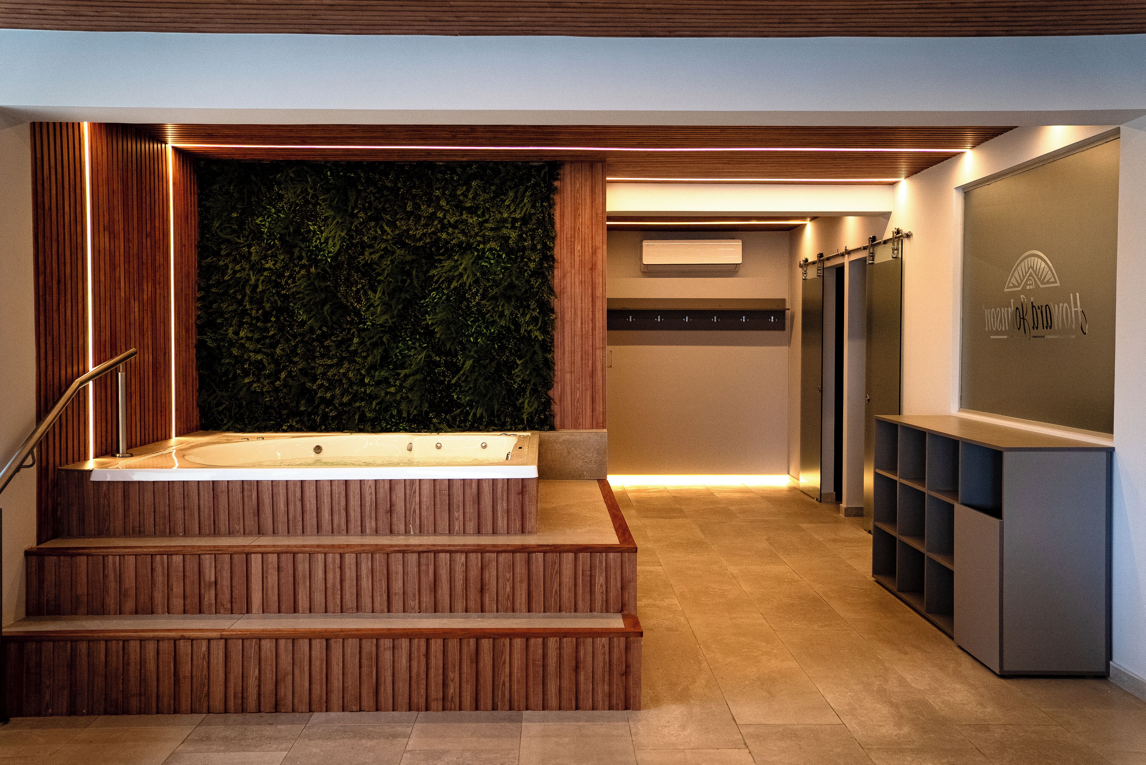 Ein modernes Interieur mit einer Holztreppe zu einem Wellnessbereich, einer Wand mit vertikalem Grün und einem Empfangstresen.