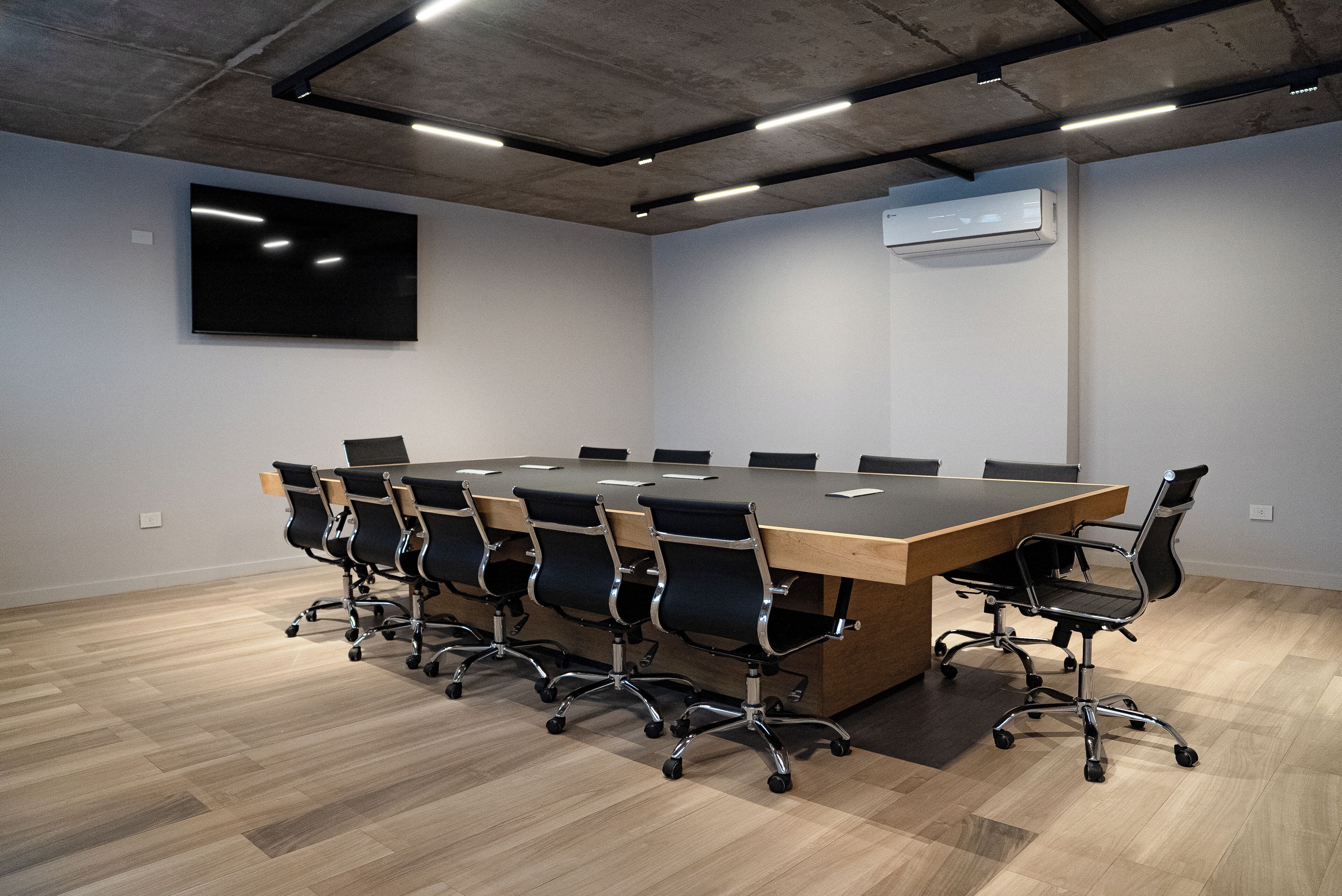 A conference room with a long table, multiple chairs, a flat-screen TV, and an air conditioning unit.