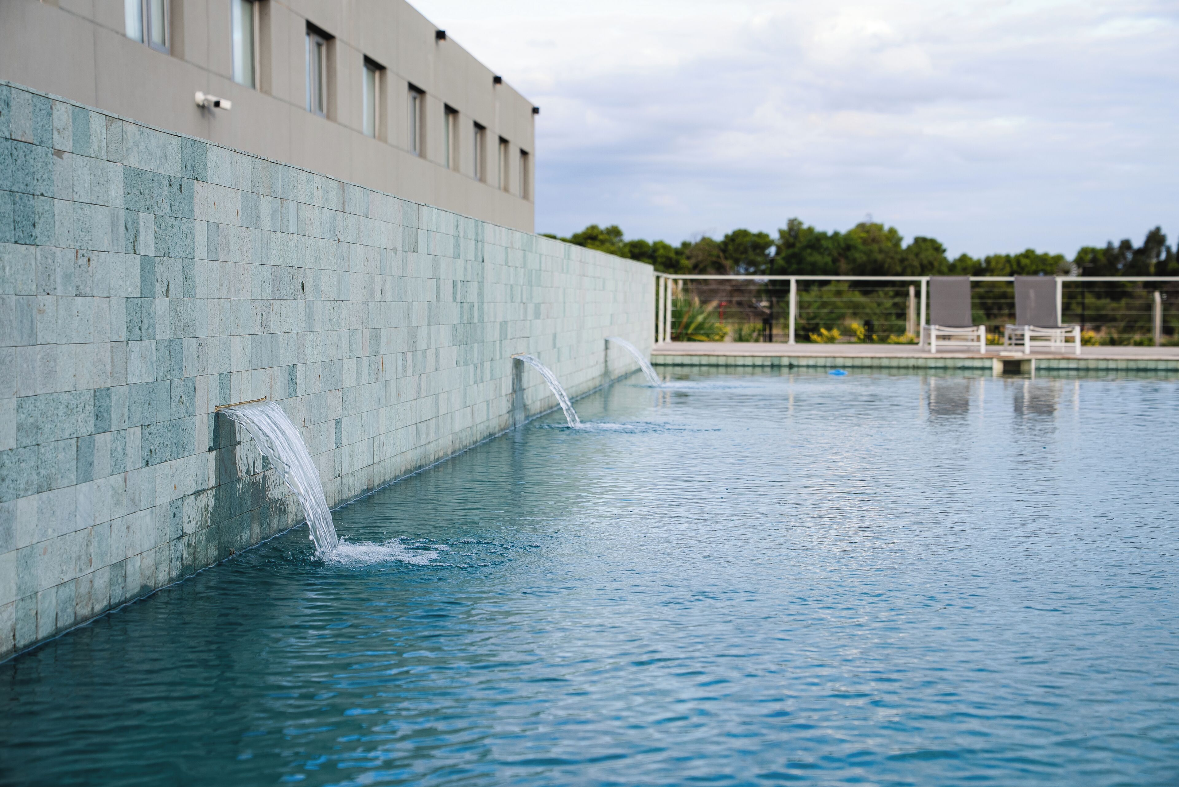 A swimming pool with water jets, a tiled wall, and a building in the background.