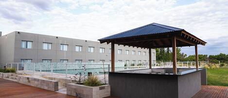 Una terraza moderna al aire libre con piso de madera, una jardinera de piedra con plantas y un pabellón con techo metálico.
