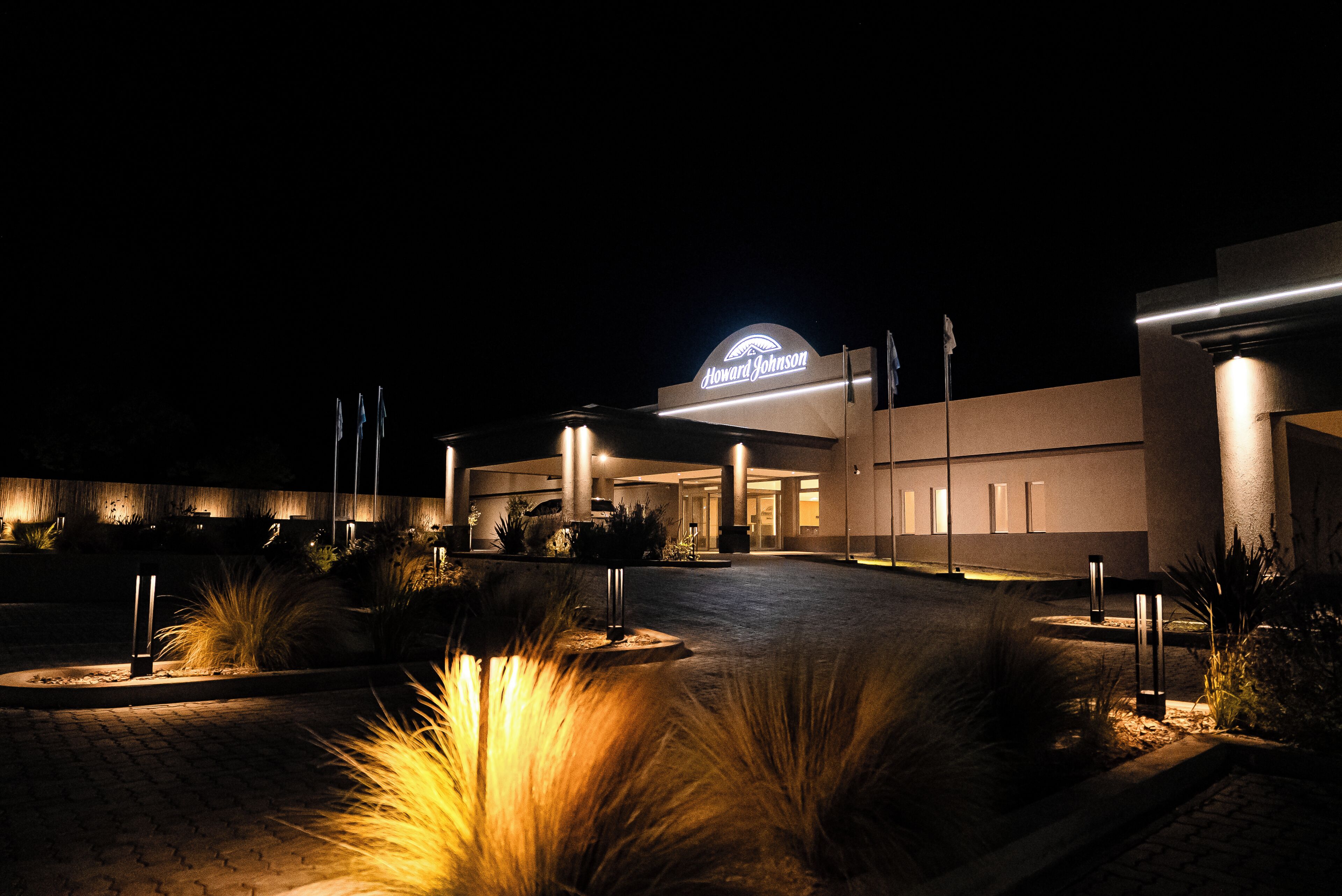 A hotel entrance at night with a well-lit sign, paved driveway, and landscaped areas with plants.