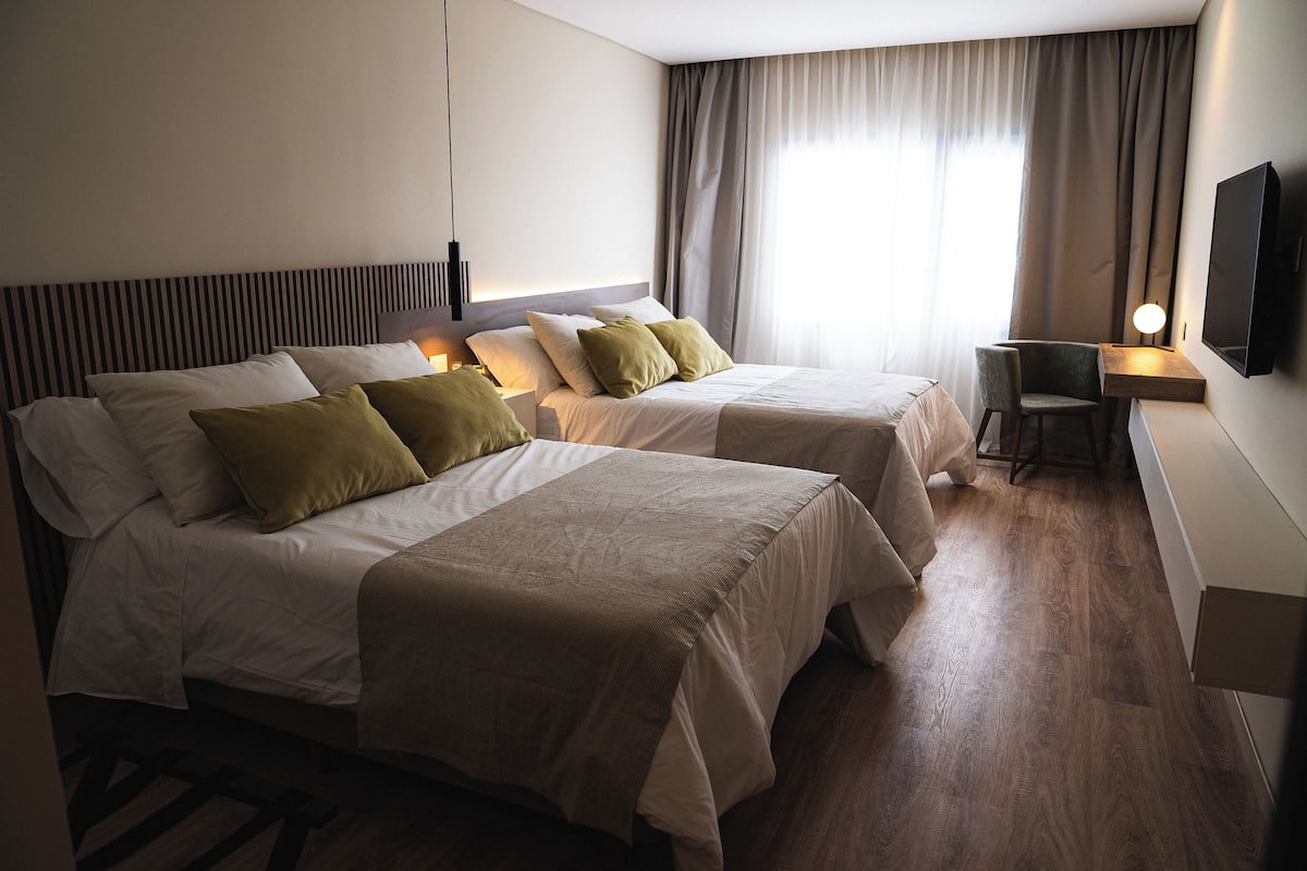 A hotel room with two beds, a desk, a chair, a TV, and a window with curtains.