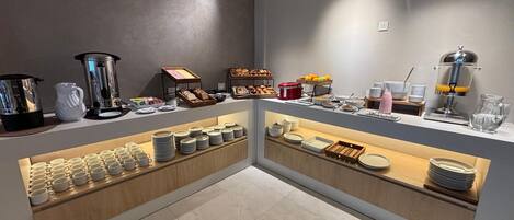 Un bufé de desayuno con diversas opciones de alimentos y bebidas, una máquina de café y una presentación de frutas.