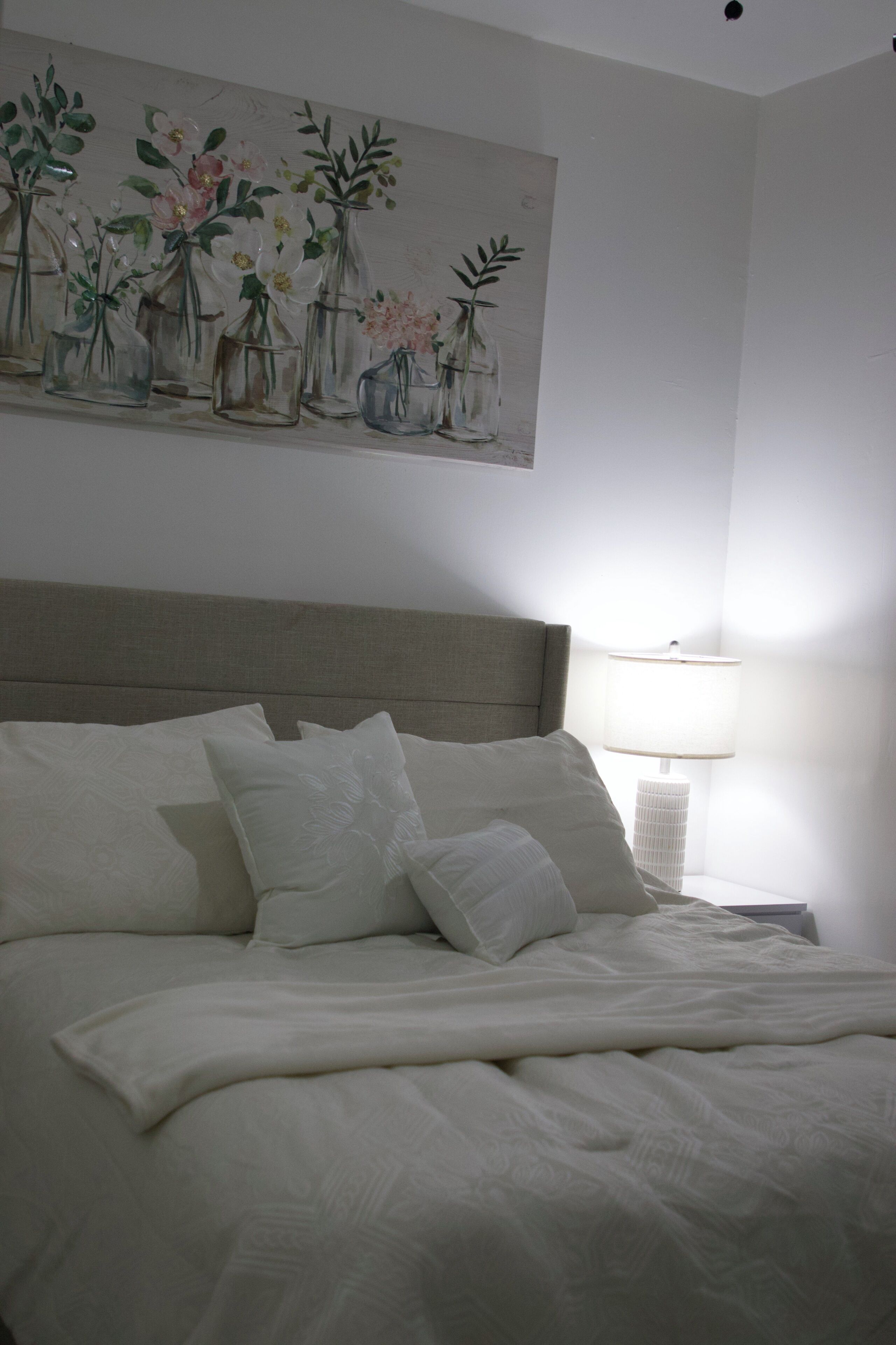 Un lit bien fait, avec du linge de lit blanc, une tête de lit et un tableau accroché au mur représentant des vases de fleurs.