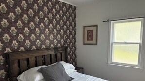 A bed with white bedding and a wooden headboard, a small bedside table, a framed picture on the wall, and a window with blinds.