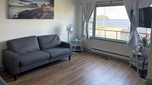 A room with a gray sofa, a ceiling fan, a painting of a lighthouse, a window with curtains, and a view of the sea.
