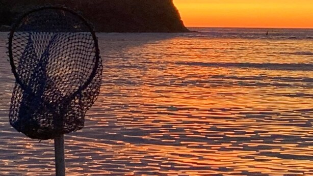 A coastal scene with a netted object in the foreground, a rocky outcrop with a structure on top, and a sunset over the water.