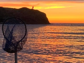 A coastal scene with a netted object in the foreground, a rocky outcrop with a structure on top, and a sunset over the water.