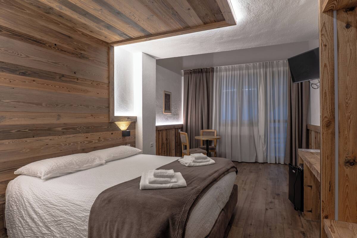 Un dormitorio con techo de madera, cama con ropa de cama blanca y marrón, escritorio con silla y televisor montado en la pared.