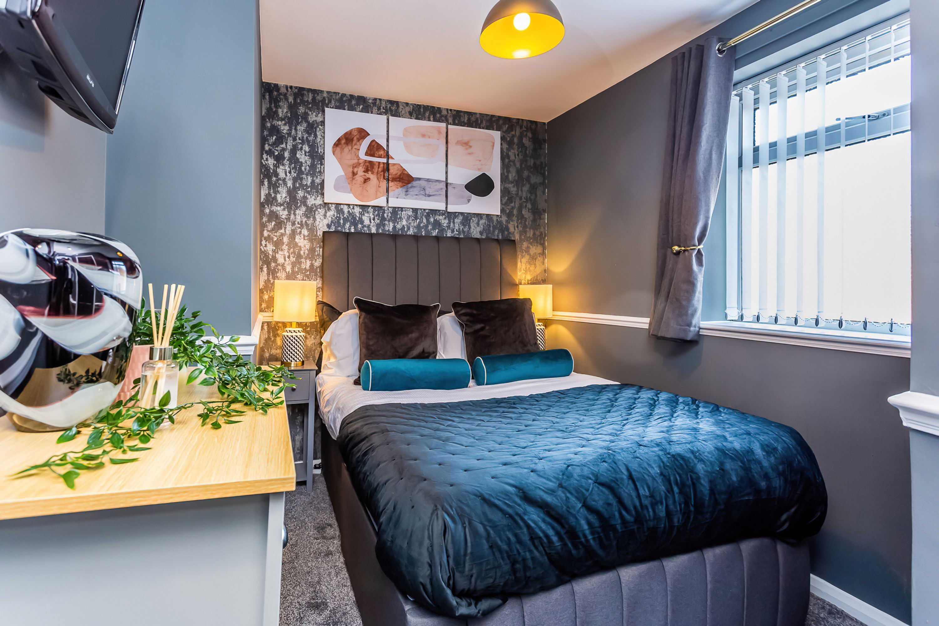 A modern bedroom with a bed, bedside table, lamp, and a window with blinds.