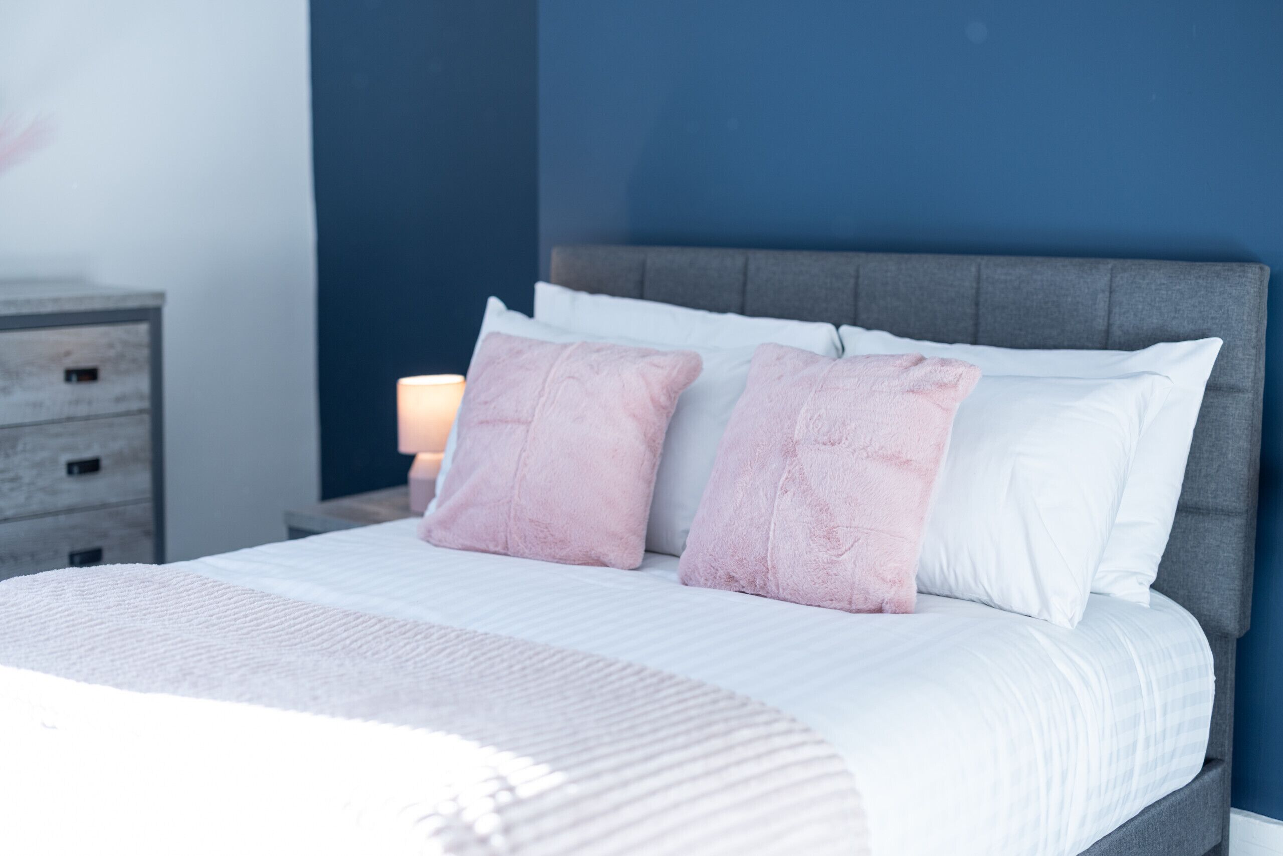 A neatly made bed with a grey headboard, white bedding, and two pink pillows.