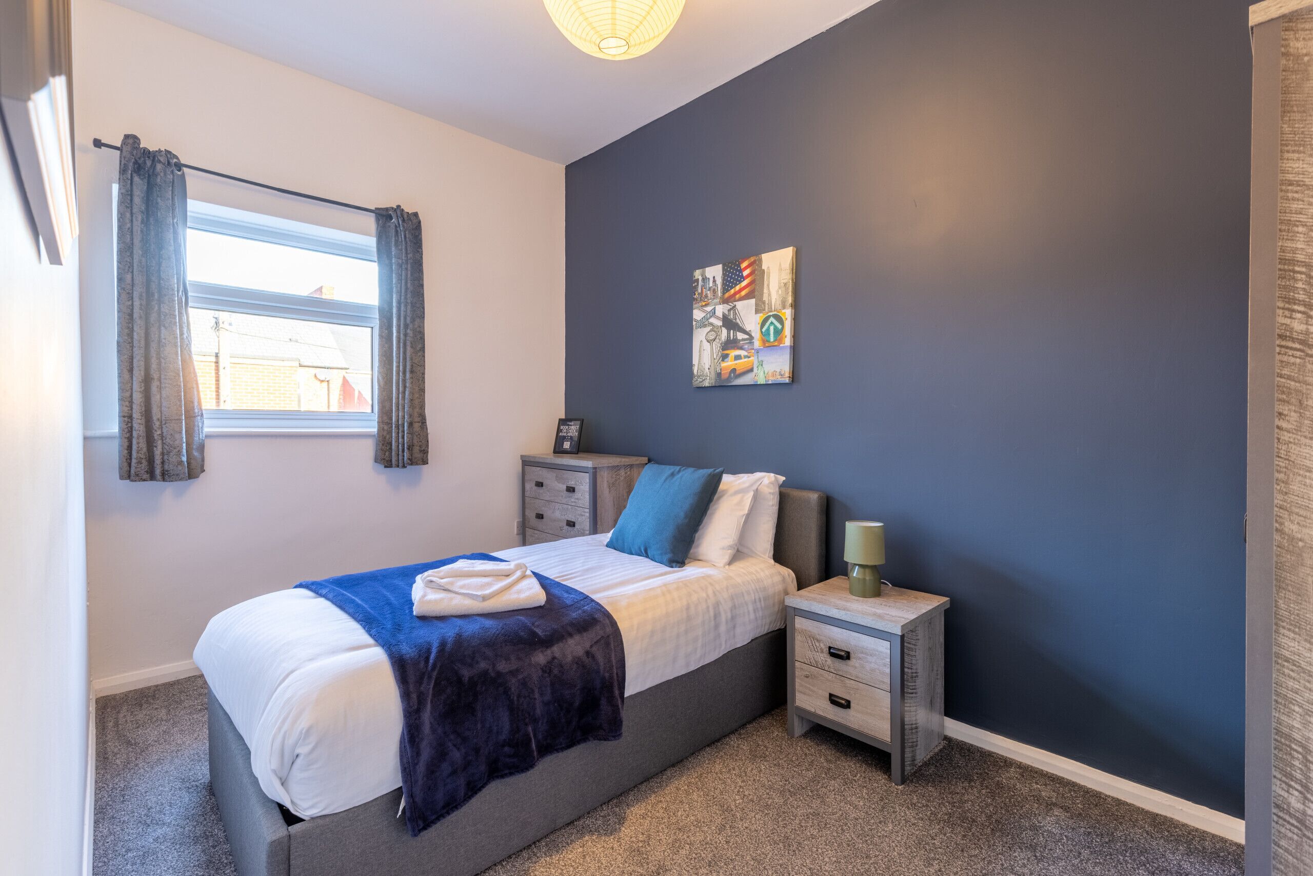 A bedroom with a bed, nightstands, a lamp, a window with curtains, and a wall with a painting.