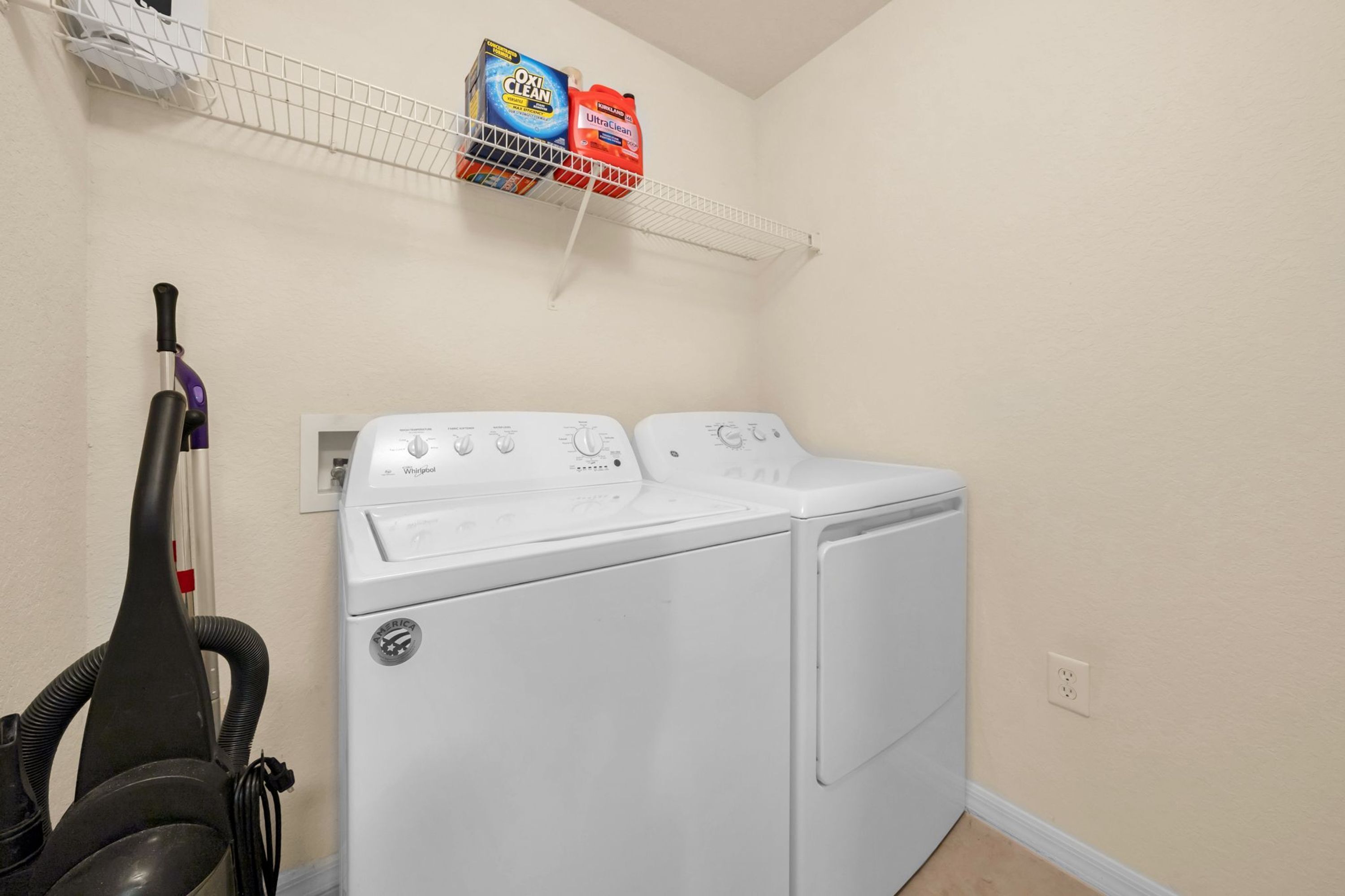 Un espace buanderie avec un lave-linge et un sèche-linge blancs, un aspirateur et du détergent rangés sur une étagère.
